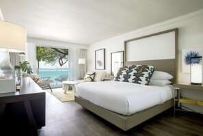 A modern hotel room with a large bed, a desk with a lamp, a chair, and a view of the ocean.