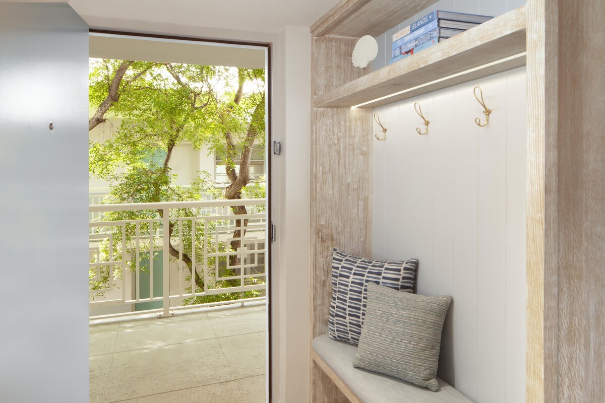 A room with a sliding glass door leading to a balcony, a bench with cushions, a wooden shelf with books, and a wall-mounted hook.