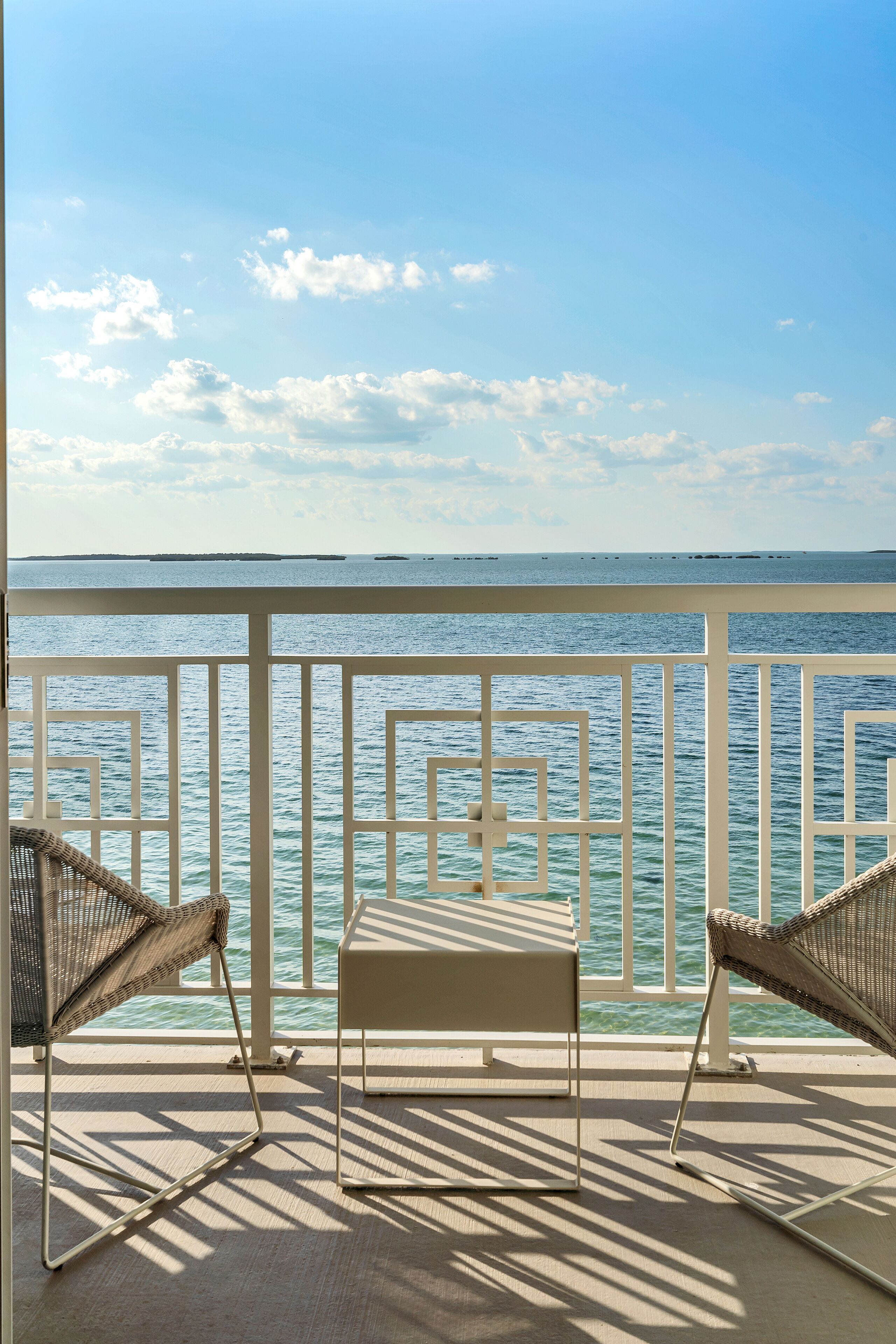 A balcony with two wicker chairs and a small table, overlooking a calm sea and distant islands.