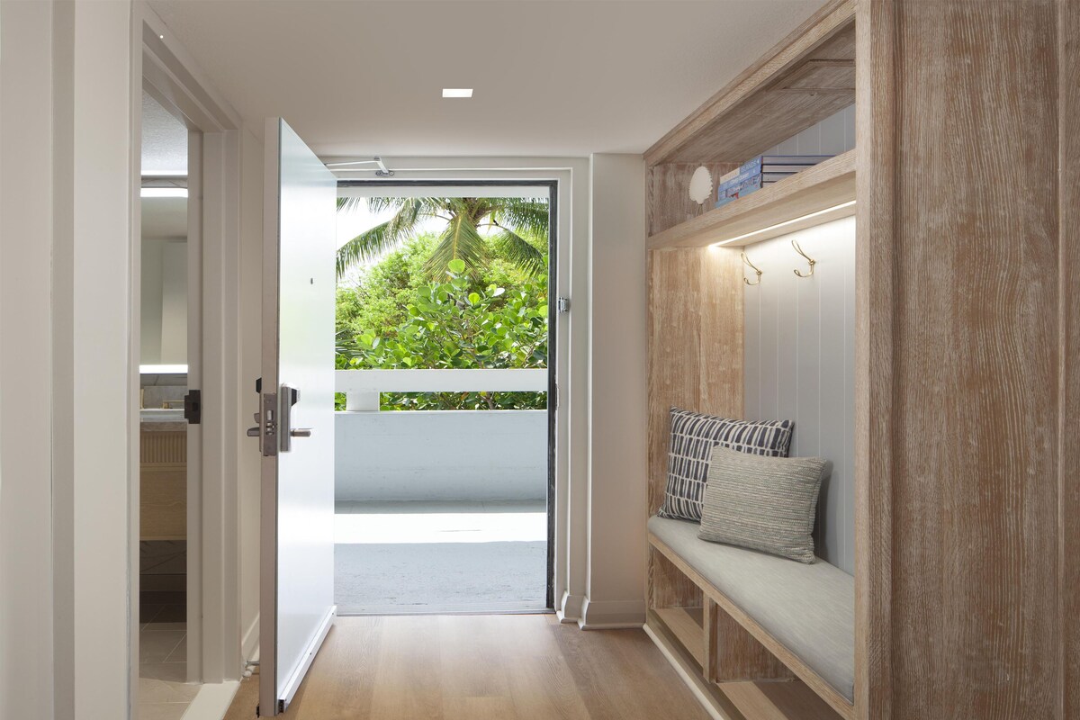 A hallway with a wooden floor, a bench with cushions, and a door leading to a balcony with greenery.