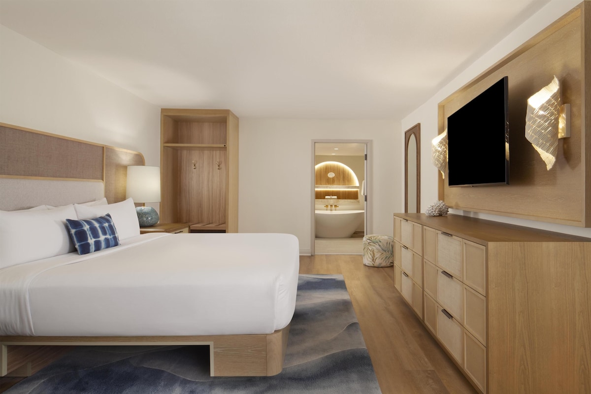 A modern hotel room with a large bed, a TV, wooden cabinets, and a bathroom visible through an open door.