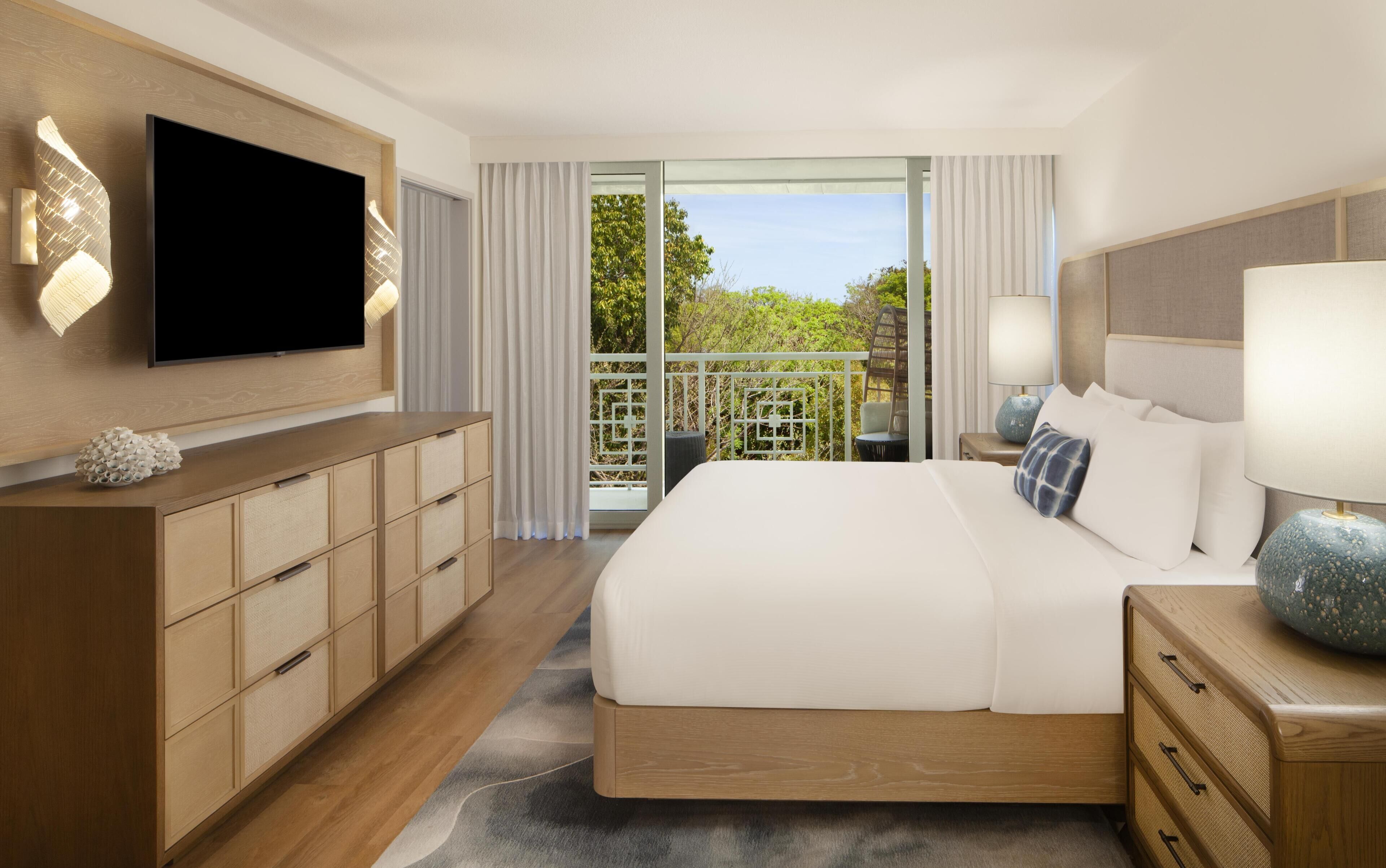 A hotel room with a large bed, a TV mounted on the wall, a wooden dresser, and a balcony with a view of greenery.