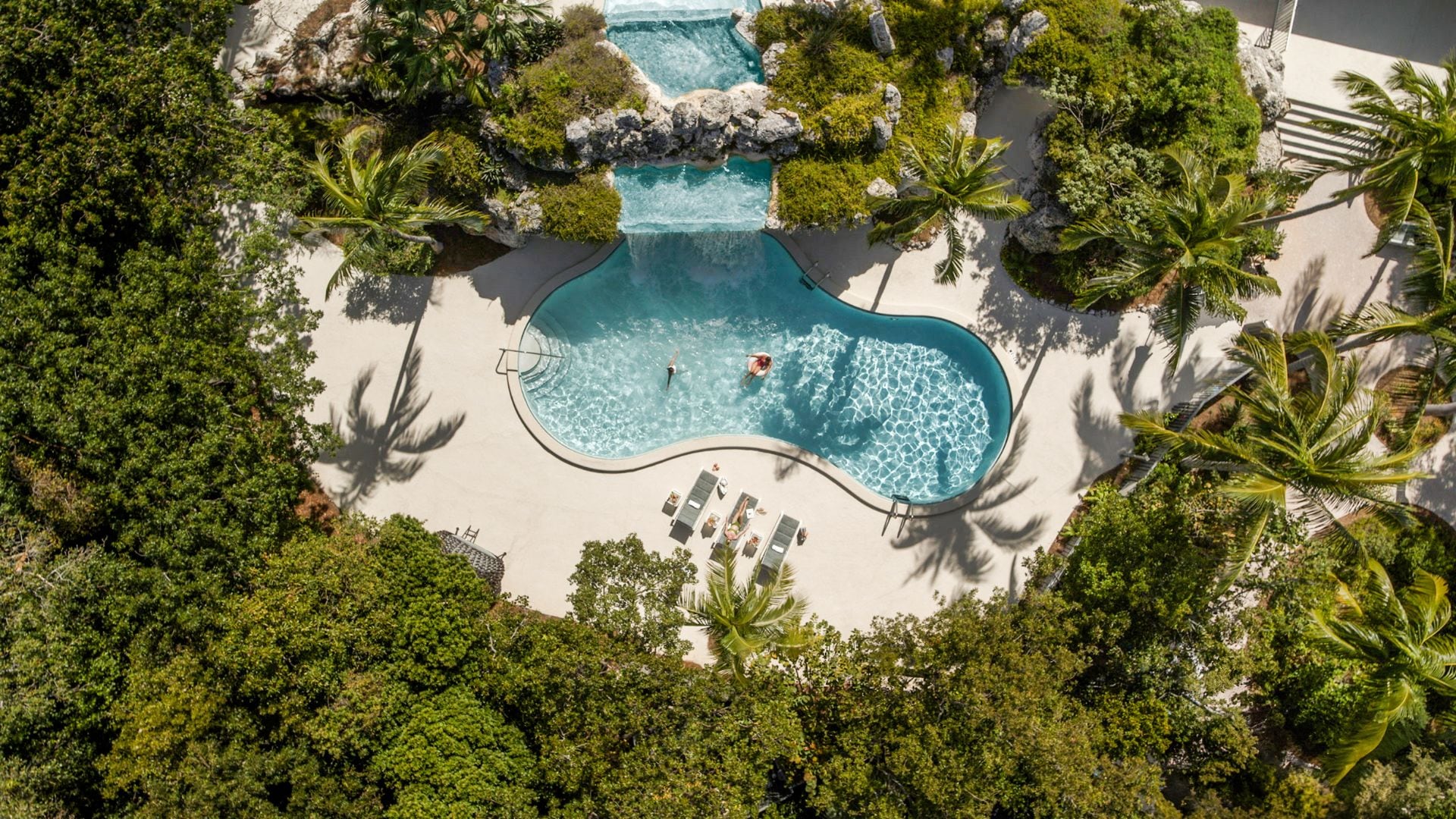An aerial view of a resort with a central swimming pool surrounded by palm trees and a sandy beach.