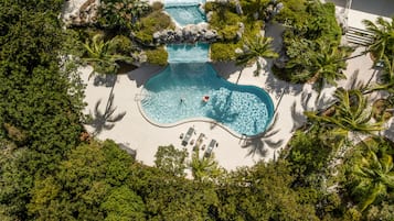 An aerial view of a resort with a central swimming pool surrounded by palm trees and a sandy beach.