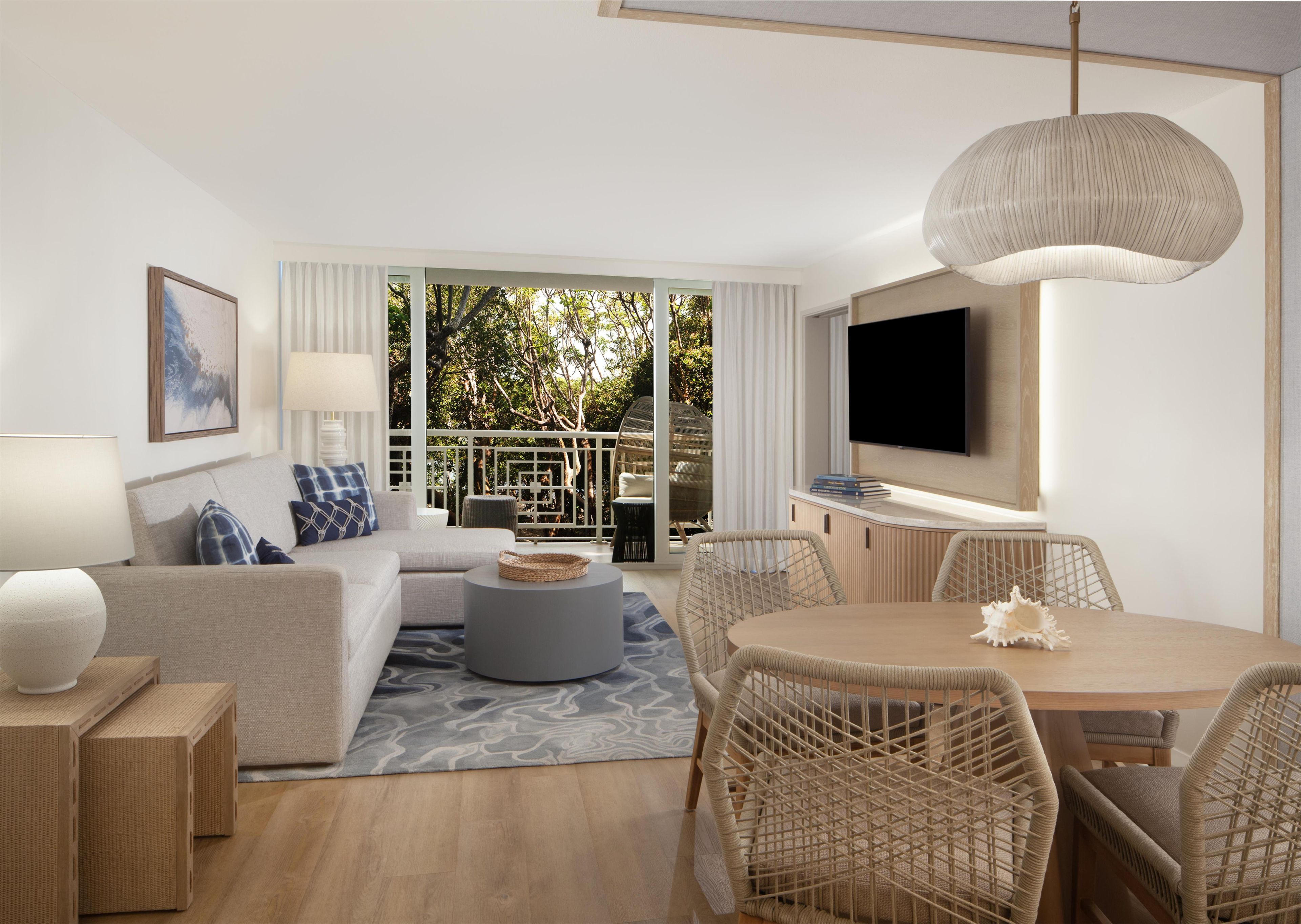 Een moderne woonkamer met een bank, salontafel, eettafel, rieten stoelen, een televisie en een balkon met uitzicht op bomen.