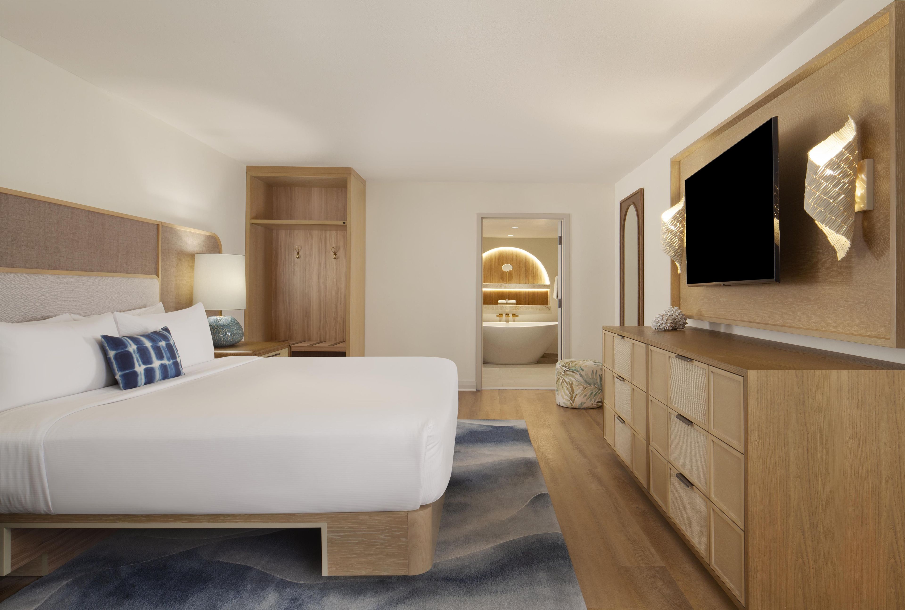 A modern hotel room with a large bed, a TV, wooden cabinets, and a bathroom visible through an open door.