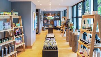 A store with wooden shelves displaying various products, a central display table with items, and a view of the outdoors through large windows.