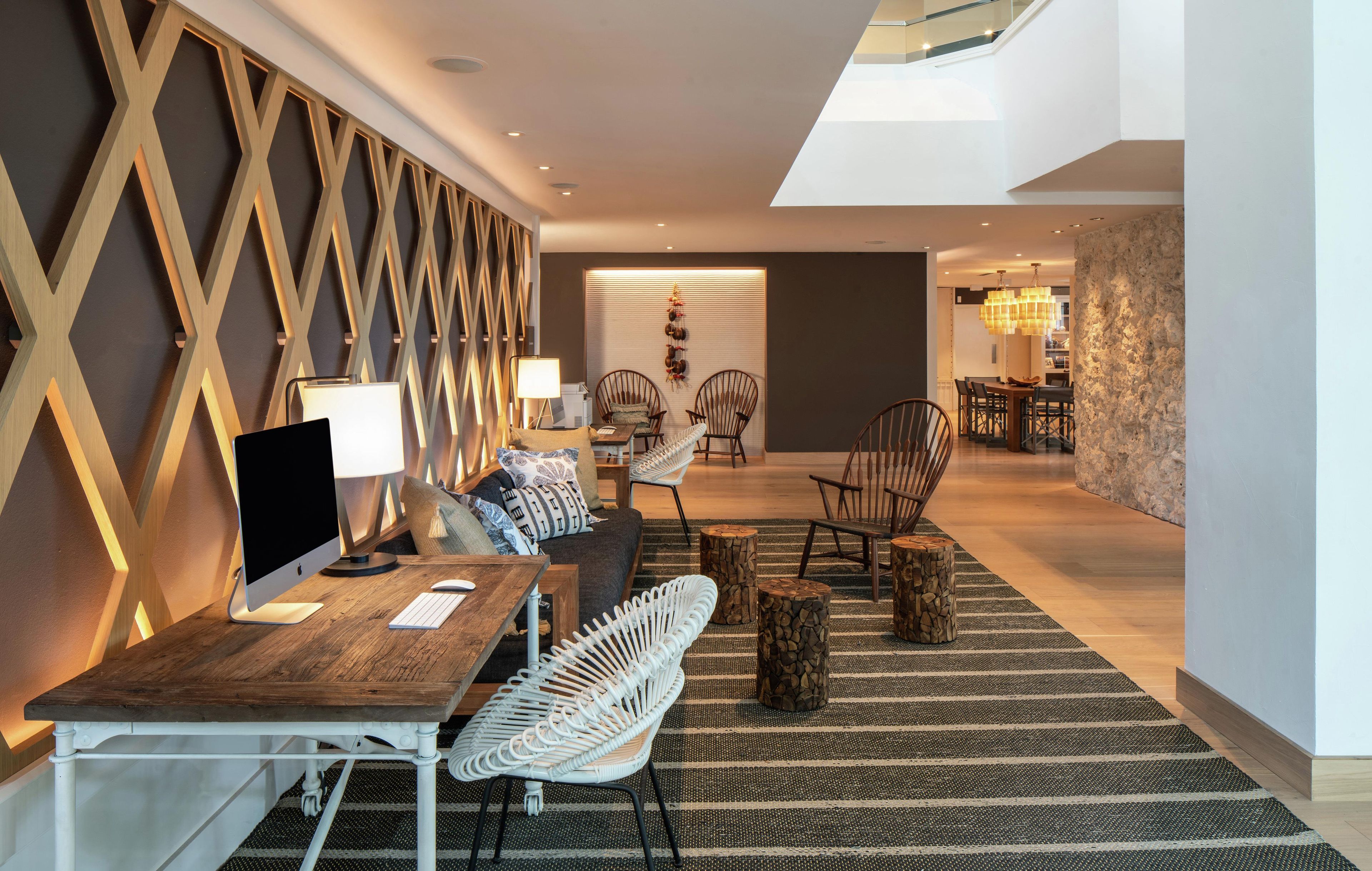 Un salon moderne avec un bureau en bois, un ordinateur et un canapé. Il y a deux fauteuils et une lampe. Le mur est décoré d’un motif et la pièce offre une vue sur une autre pièce où se trouve une table à manger.