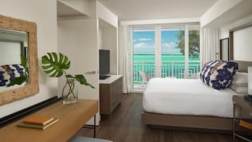 A hotel room with a bed, a desk, a chair, a TV, a plant, and a view of the ocean.