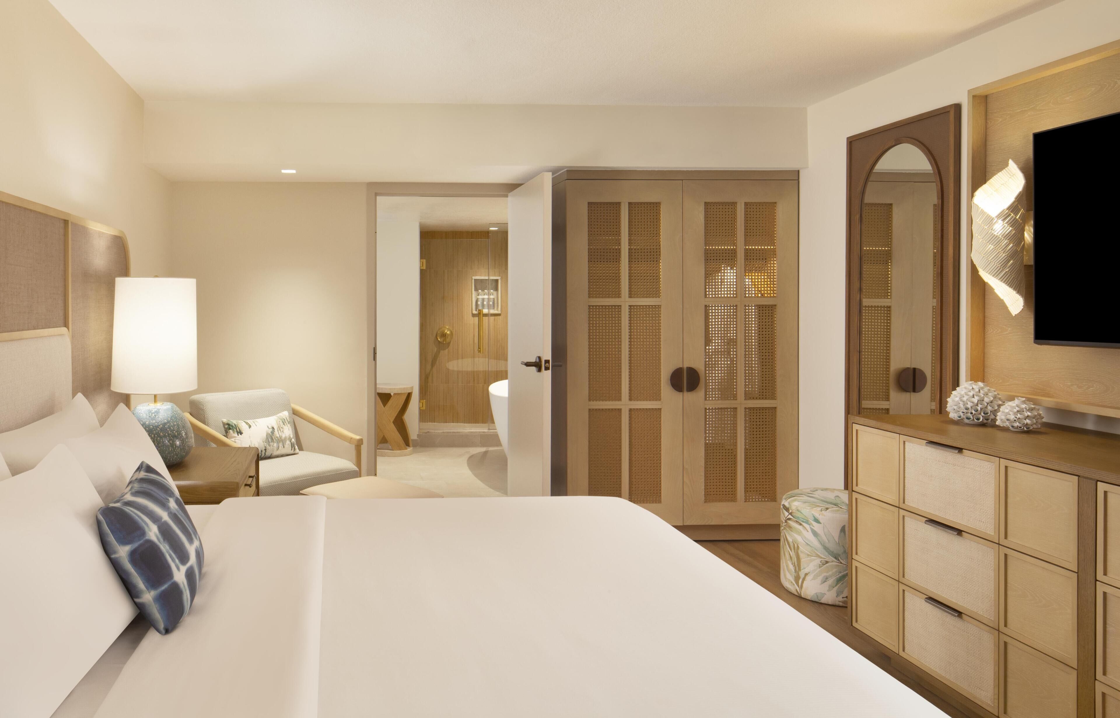 A hotel room with a large bed, a desk, a chair, a wardrobe, a television, and a bathroom visible through an open door.