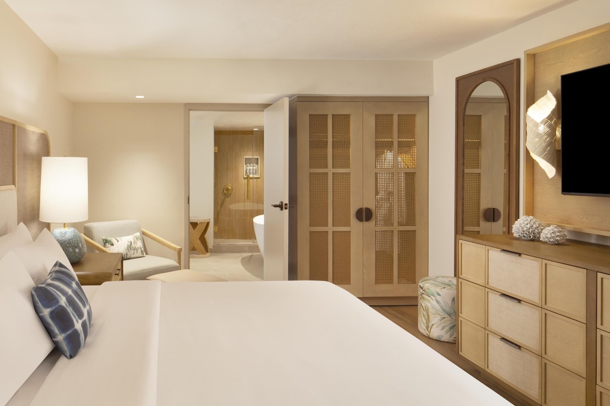 A hotel room with a large bed, a desk, a chair, a wardrobe, a television, and a bathroom visible through an open door.
