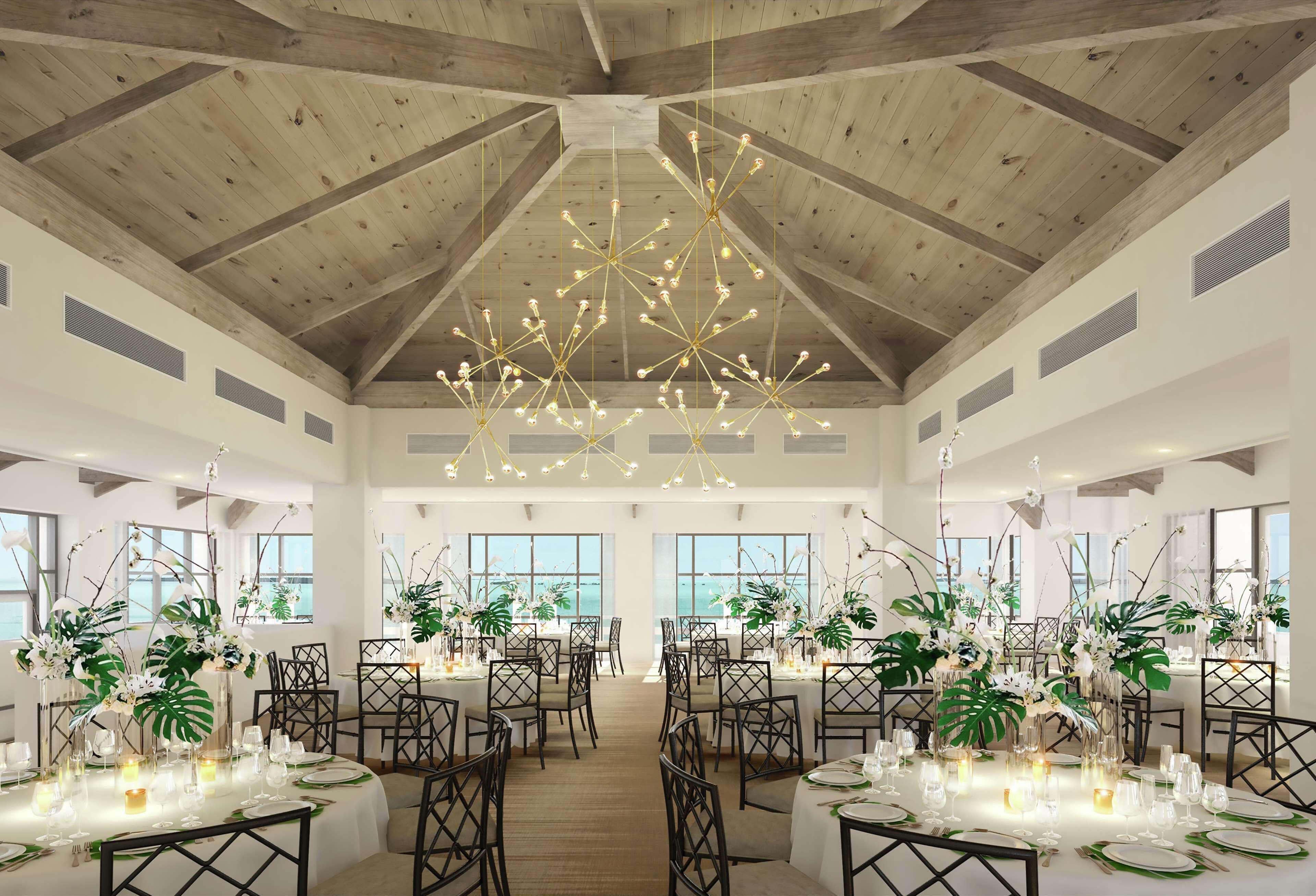 A spacious dining area with high ceilings and wooden beams, featuring tables set with white tablecloths, green centerpieces, and candles.