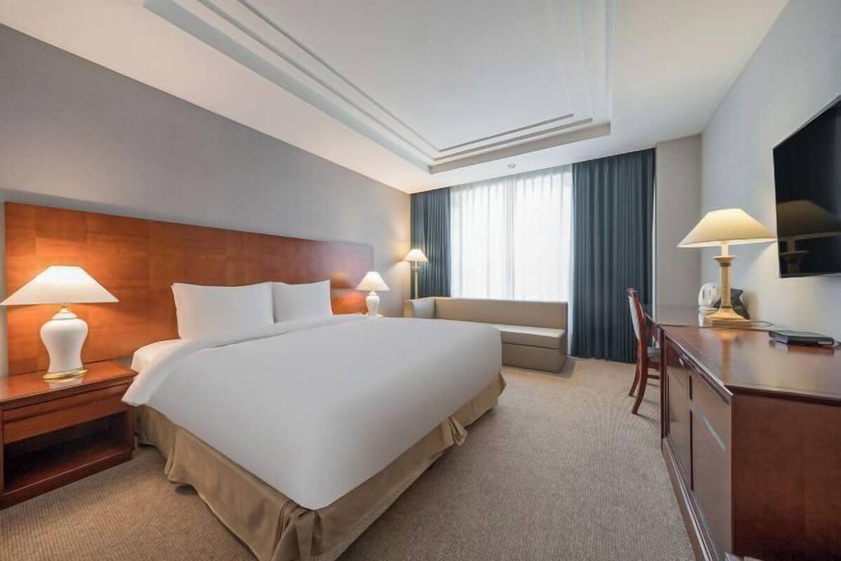 Ein Hotelzimmer mit einem großen Bett, zwei Nachttischen mit Lampen, einem Sofa, einem Schreibtisch mit Stuhl und einem an der Wand befestigten Fernseher.