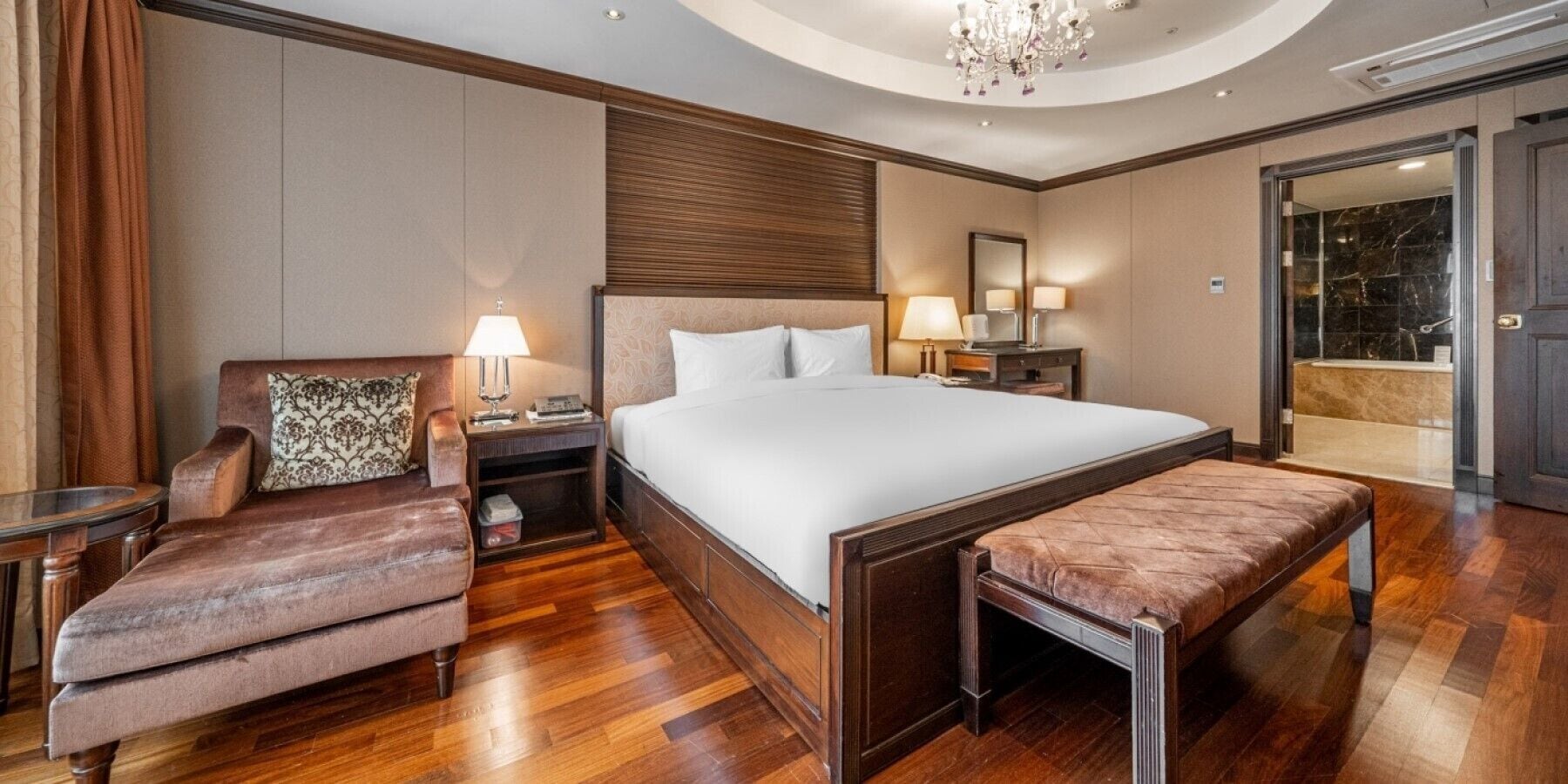 Habitación de hotel con una cama grande, una zona de estar con una silla y un banco, una mesita de noche con lámpara, y un baño visible a través de una puerta abierta.