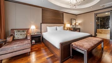 Habitación de hotel con una cama grande, una zona de estar con una silla y un banco, una mesita de noche con lámpara, y un baño visible a través de una puerta abierta.