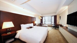 A hotel room with a large bed, a desk, a chair, a TV, and a window with a city view. - Koreana Hotel (Seoul)