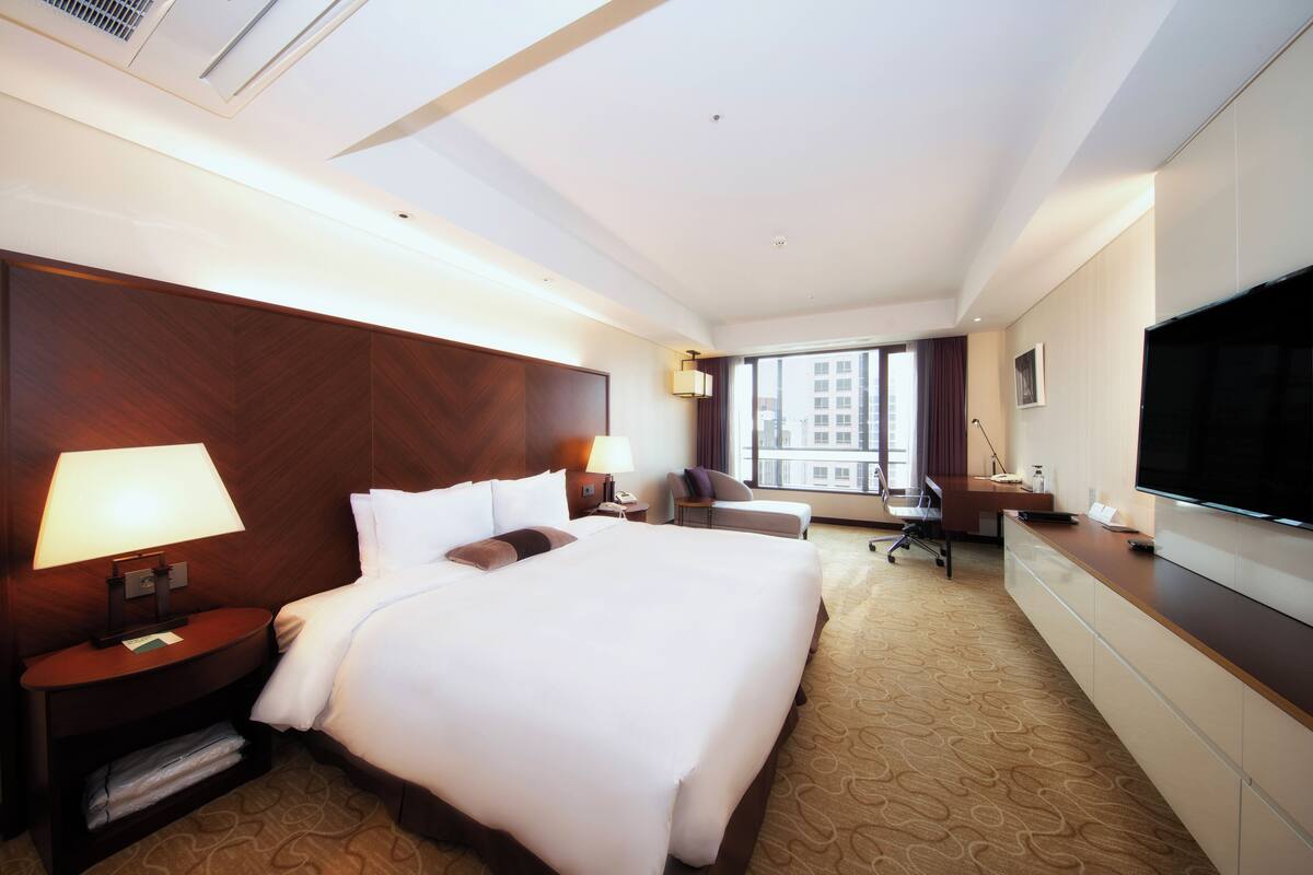 Ein Hotelzimmer mit einem großen Bett, einem Schreibtisch, einem Sessel, einem Fernseher und einem Fenster mit Blick auf die Stadt.