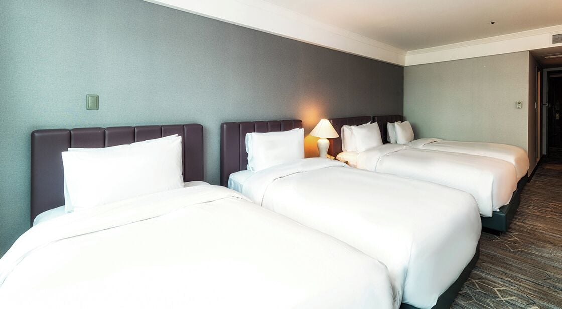 Habitación de hotel con tres camas, una lámpara sobre la cama central y una puerta a la derecha.