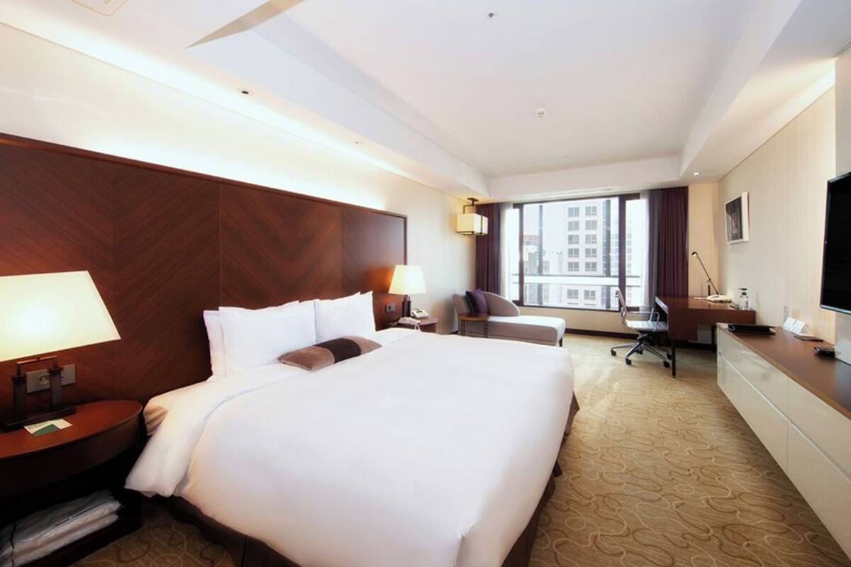 Ein Hotelzimmer mit einem großen Bett, einem Schreibtisch, einem Stuhl, einem Fernseher und einem Fenster mit Blick auf die Stadt.