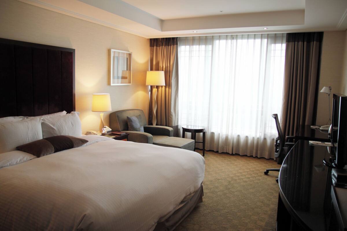 Ein Hotelzimmer mit einem großen Bett, einem Schreibtisch, einem Sessel, einem Fernseher und einem Fenster mit Vorhängen.
