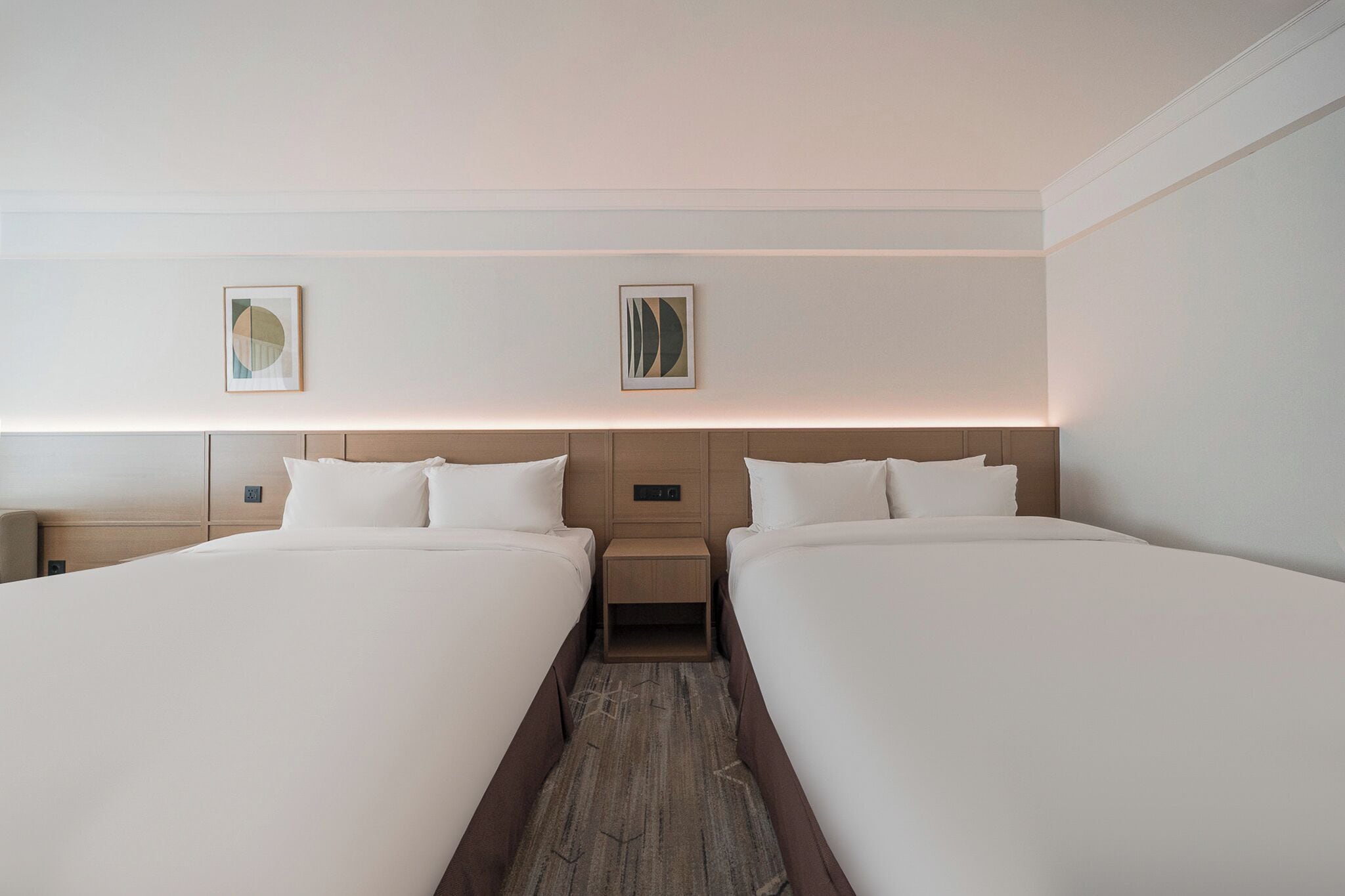 A hotel room with two beds, a nightstand, and two framed pictures on the wall.