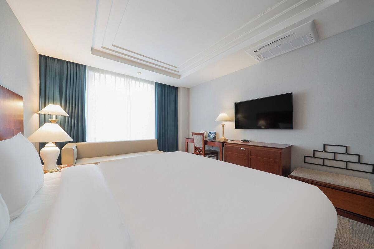 Ein Hotelzimmer mit einem großen Bett, einem Schreibtisch mit Stuhl, einem an der Wand montierten Flachbildfernseher und einem Vorhangfenster.