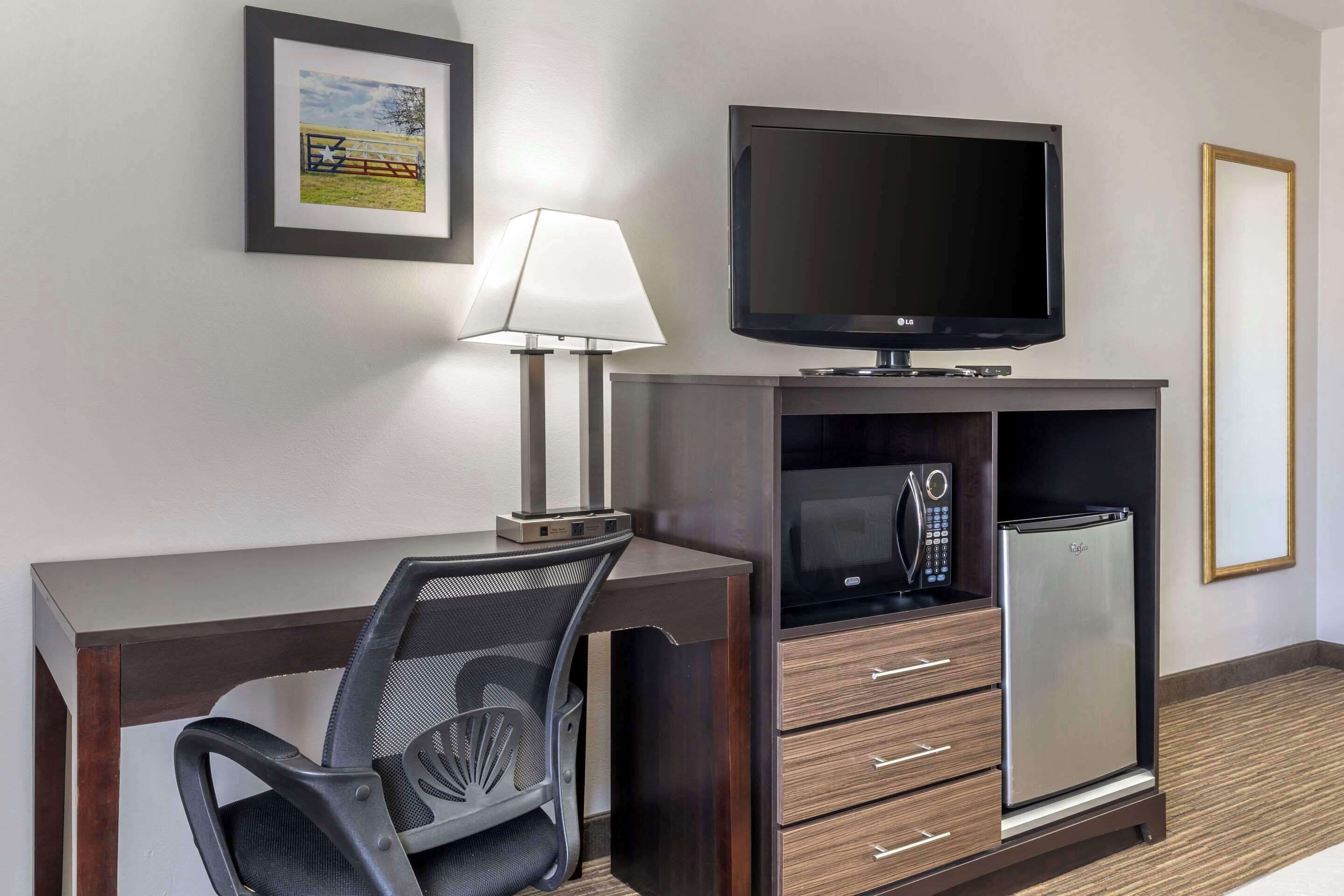 A hotel room with a desk, chair, TV, microwave, and refrigerator.