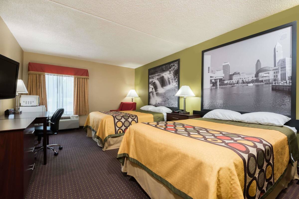Ein Hotelzimmer mit zwei Betten, einem Schreibtisch, einem Sessel, einem Fernseher und einem großen Stadtbild an der Wand.