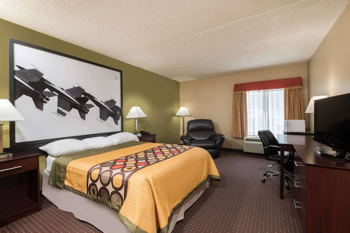 Ein Hotelzimmer mit einem Bett, zwei Nachttischlampen, einem Schreibtisch mit Fernseher, einem Sessel und einem großen, gerahmten Bild an der Wand.