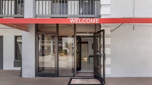 Entrada de un edificio con un cartel que dice 'WELCOME', puertas de vidrio y un pequeño espacio exterior con un felpudo.
