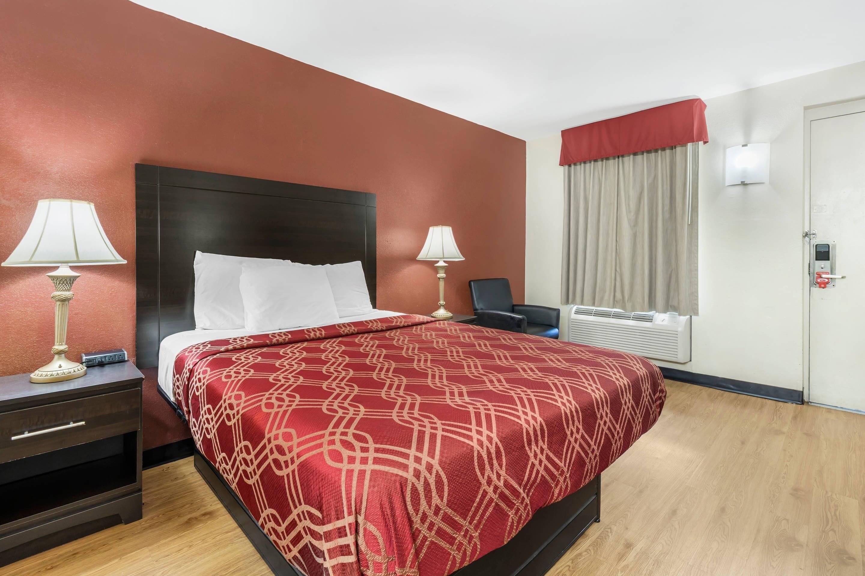 Habitación de hotel con cama, dos lámparas de noche, mesita de noche, una silla, ventana con cortinas y termostato.