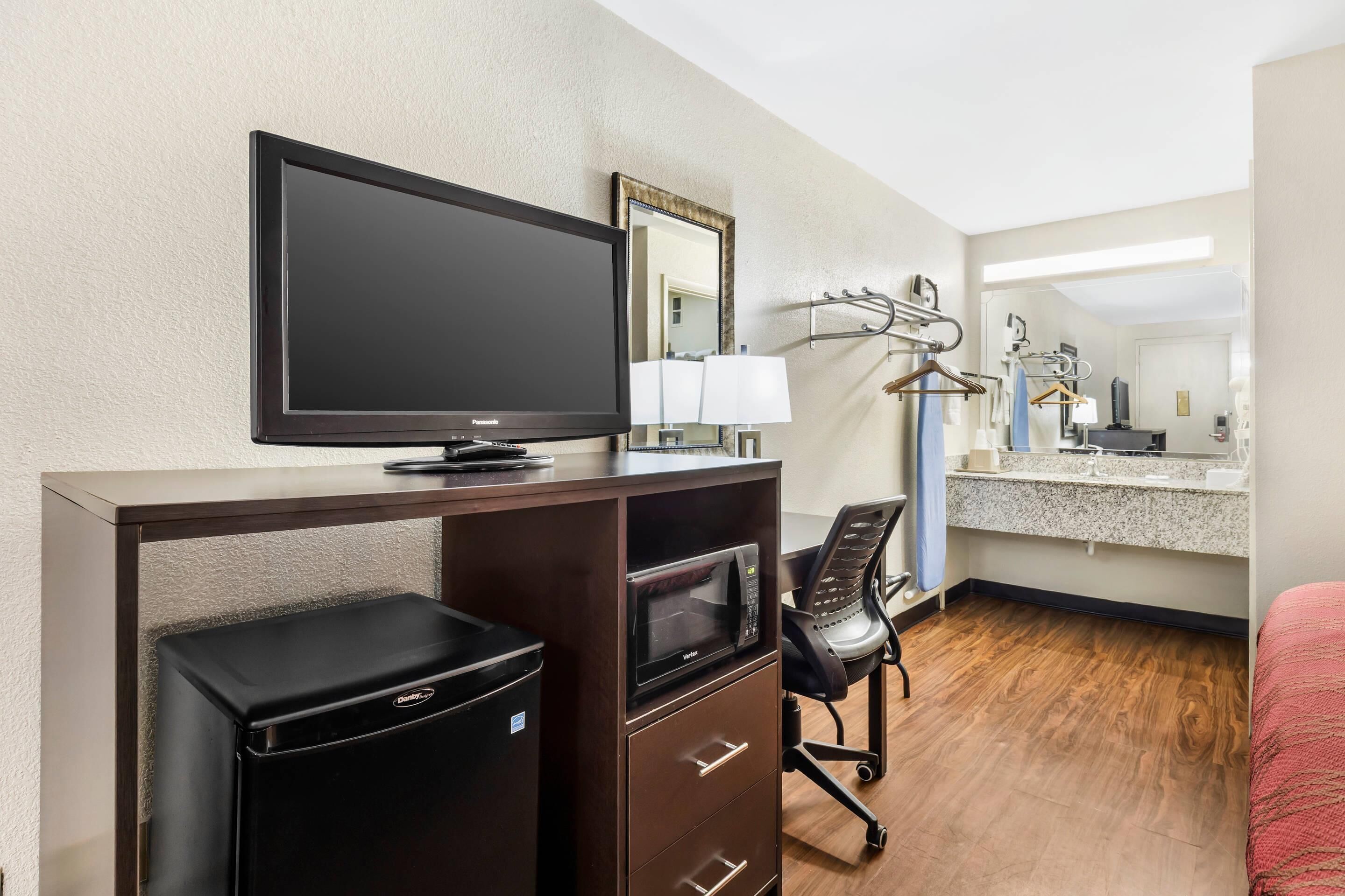 A hotel room with a desk, a television, a microwave, a chair, a bed, a bathroom with a sink and mirror, and a coat rack.