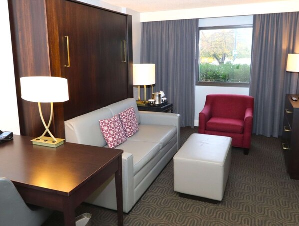 Habitación de hotel con sofá, sillón rojo, mesita, lámpara y ventana con cortinas.