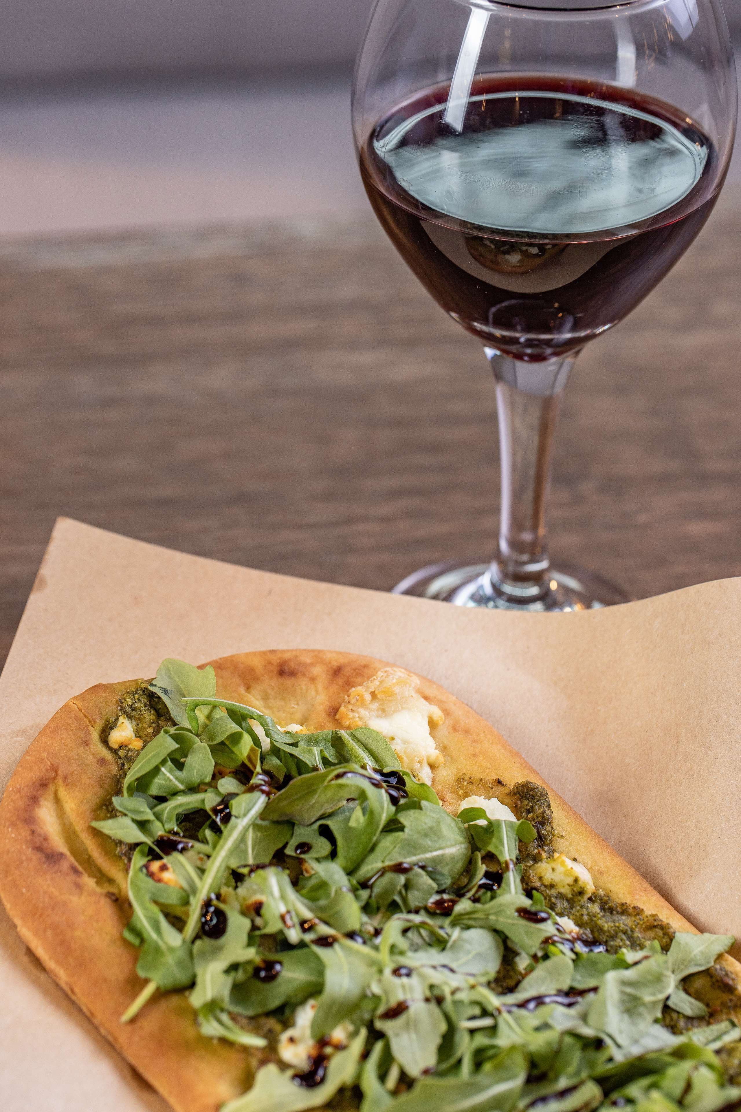 A flatbread topped with greens and cheese, accompanied by a glass of red wine on a table.