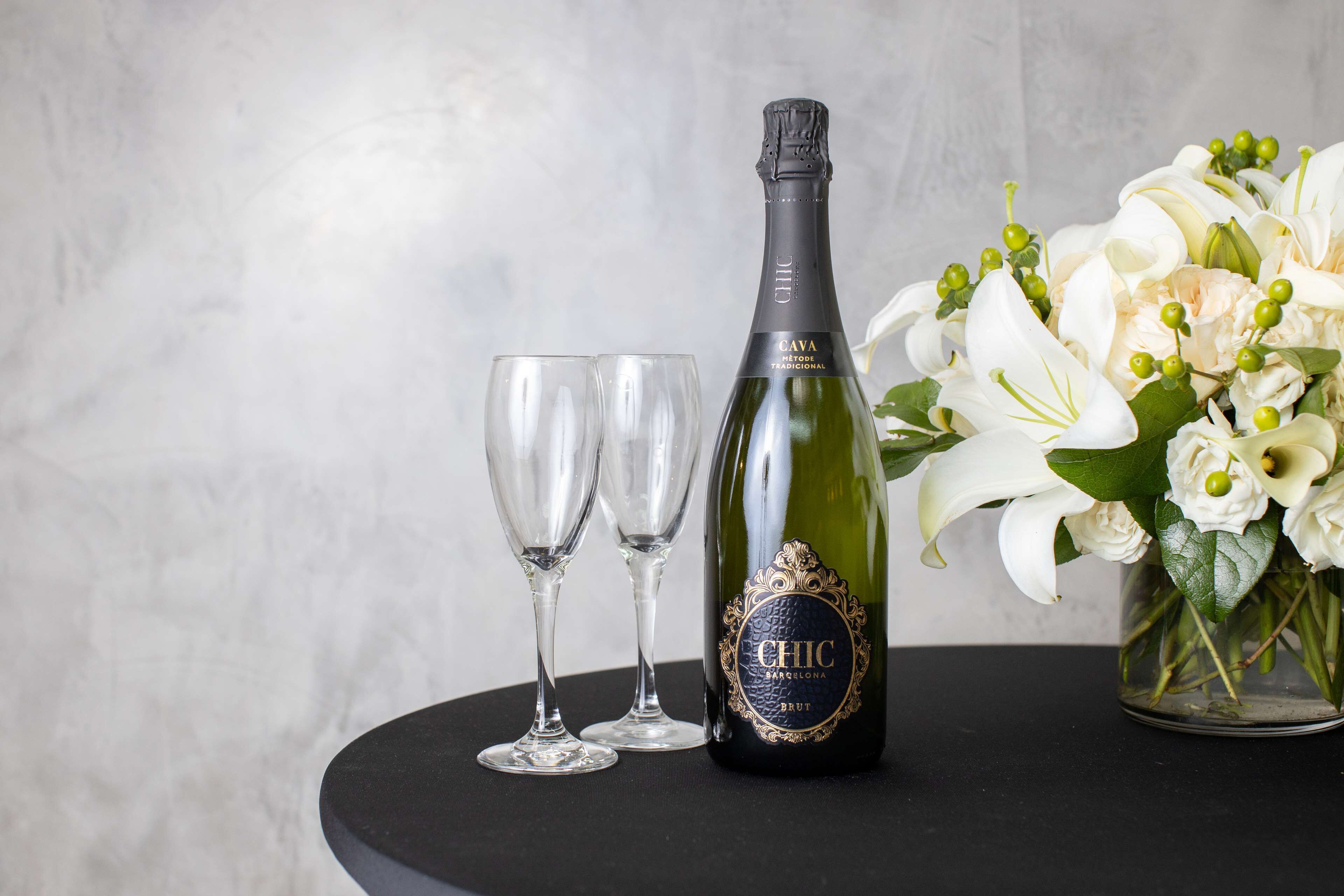 A bottle of CHIC Cava, two empty glasses, and a vase of white flowers on a black surface.