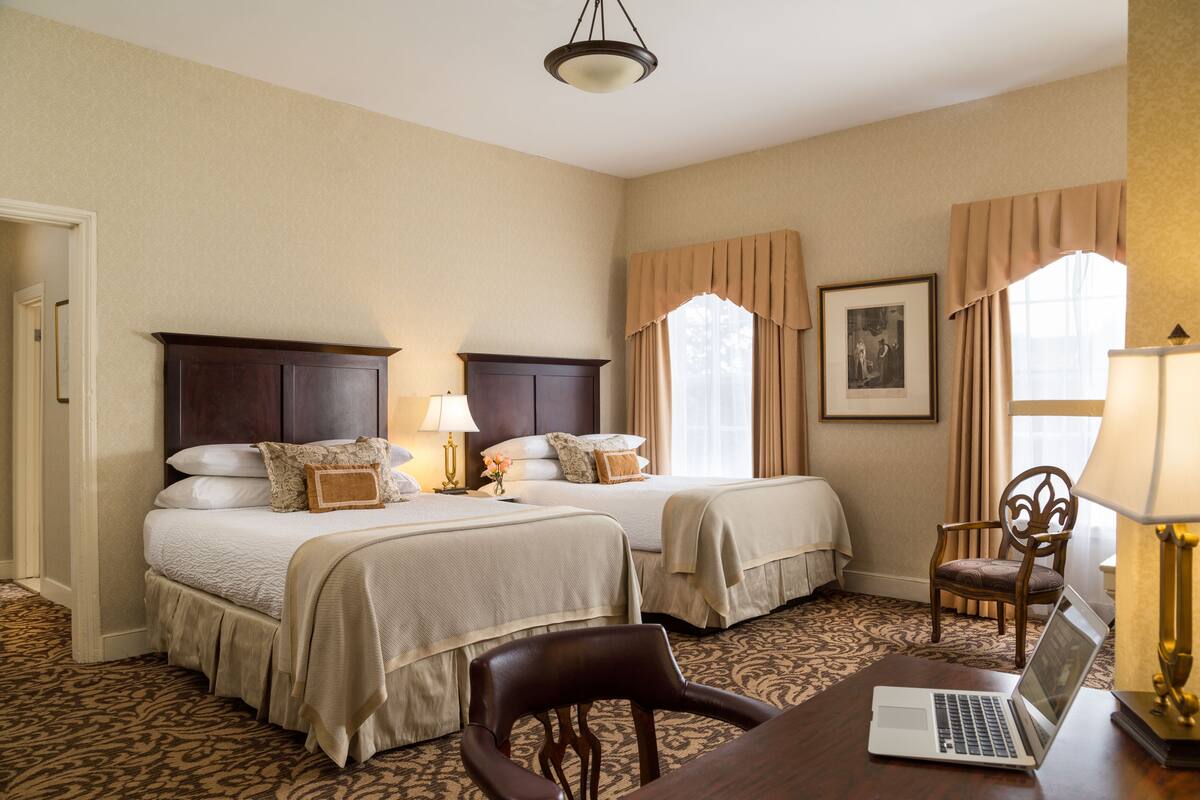A hotel room with two beds, a desk with a laptop, a chair, and a lamp.