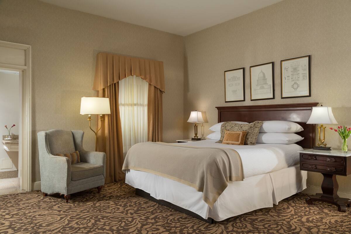 A hotel room with a large bed, two bedside lamps, a chair, a nightstand, and framed pictures on the wall.