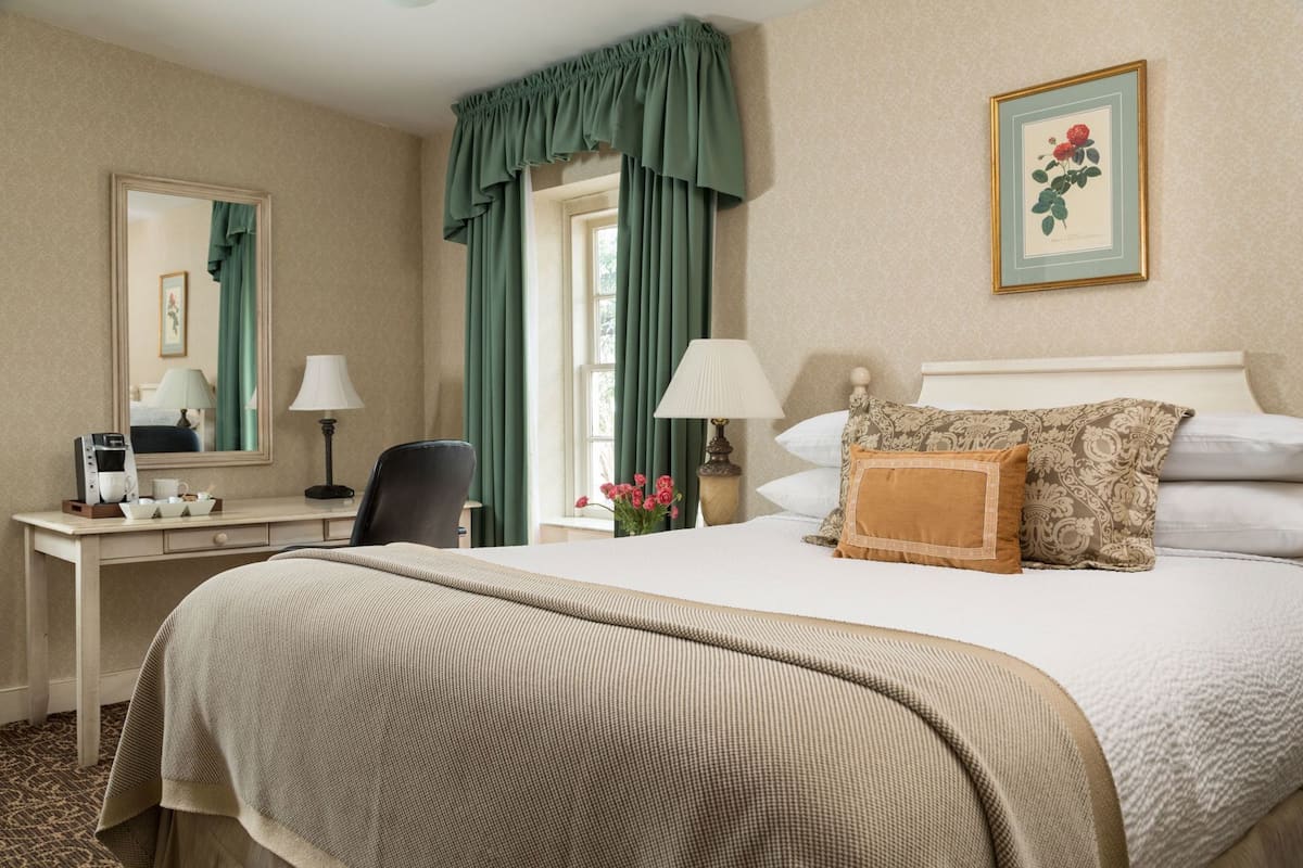 A hotel room with a bed, a desk with a chair, a lamp, a mirror, and a framed picture on the wall.