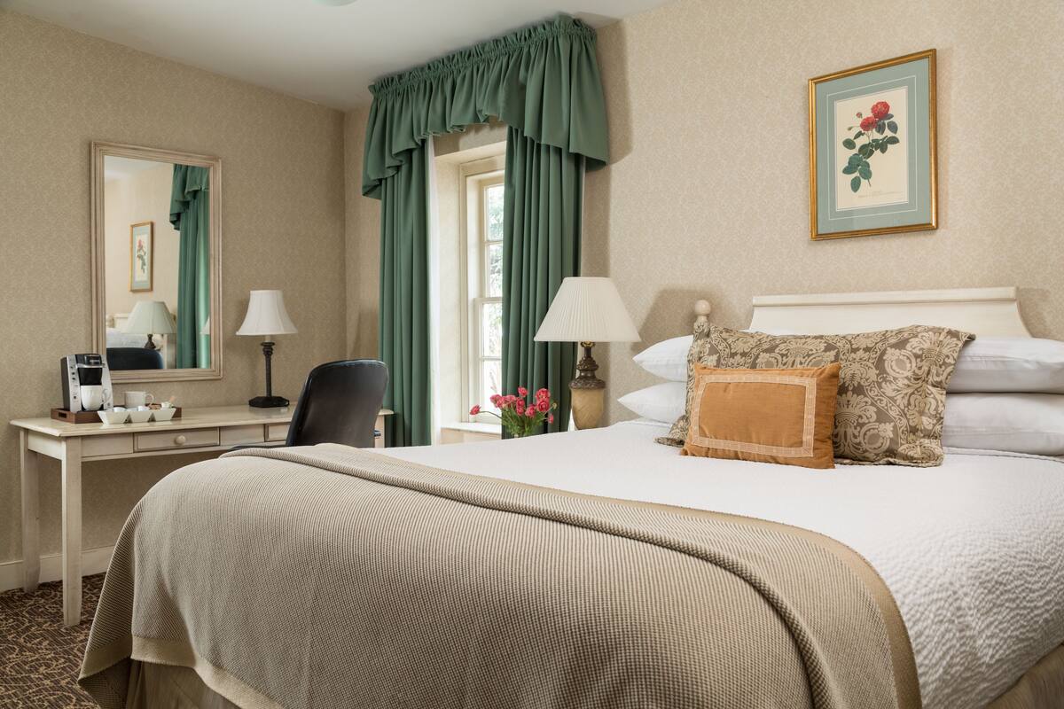 A hotel room with a large bed, a desk with a telephone and a lamp, a chair, and a window with curtains.