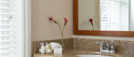 Une salle de bain avec un comptoir en granit, un lavabo blanc et un miroir au-dessus. On y trouve deux vases avec des fleurs et une serviette posée sur le comptoir.