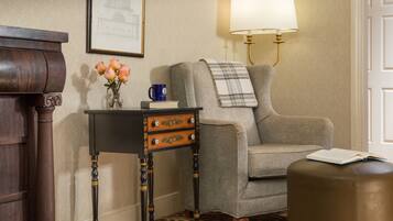 A room with a wooden sideboard, a small table with drawers, a cushioned armchair, a footstool, a lamp, and a framed picture on the wall.