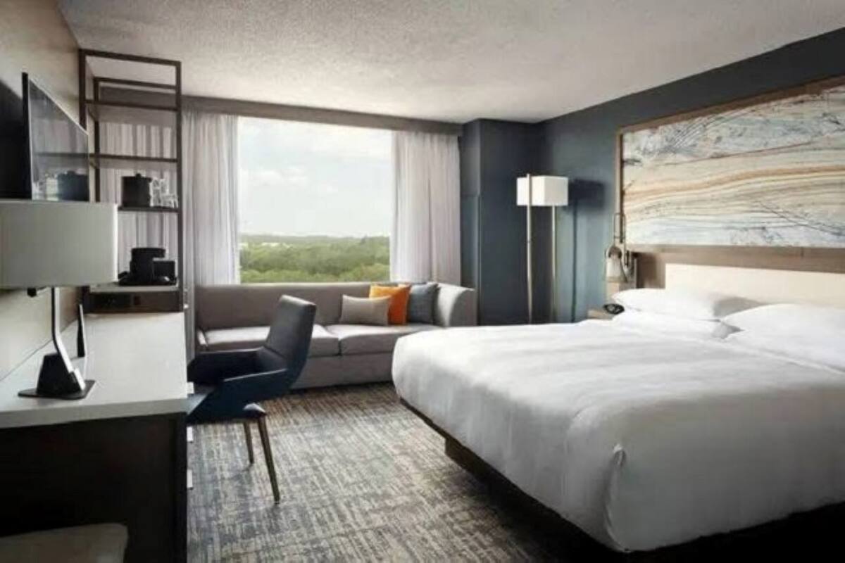 A hotel room with a bed, a desk, a chair, a sofa, a lamp, and a view of greenery.