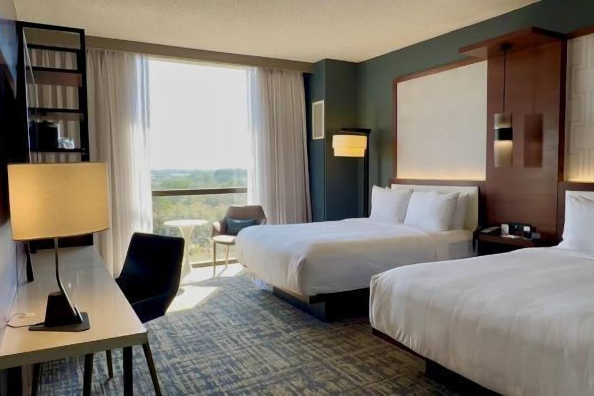 A hotel room with two beds, a desk with a lamp, a chair, and a balcony with a view.