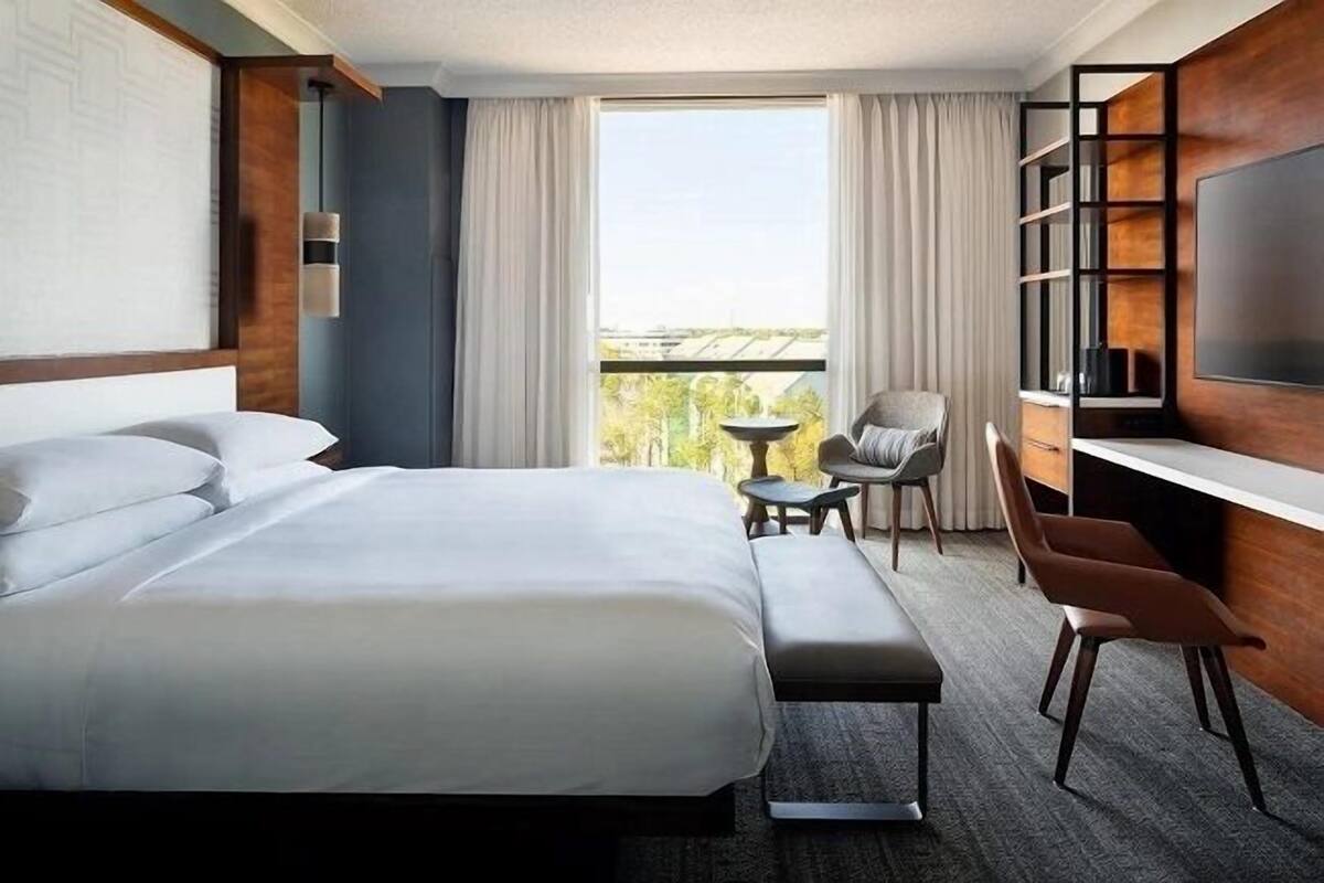 A hotel room with a large bed, a desk, a chair, a TV, and a view of greenery outside.
