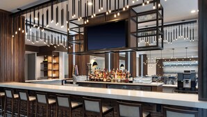 Una zona de bar moderna con una barra alargada, varios taburetes y una gran variedad de botellas y vasos.