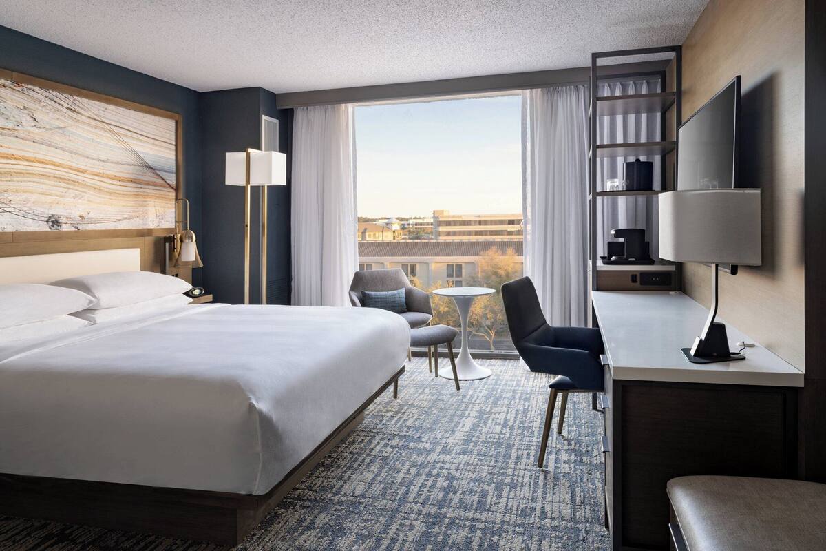 A hotel room with a large bed, a desk with a chair, a TV, a coffee maker, and a view of the city.