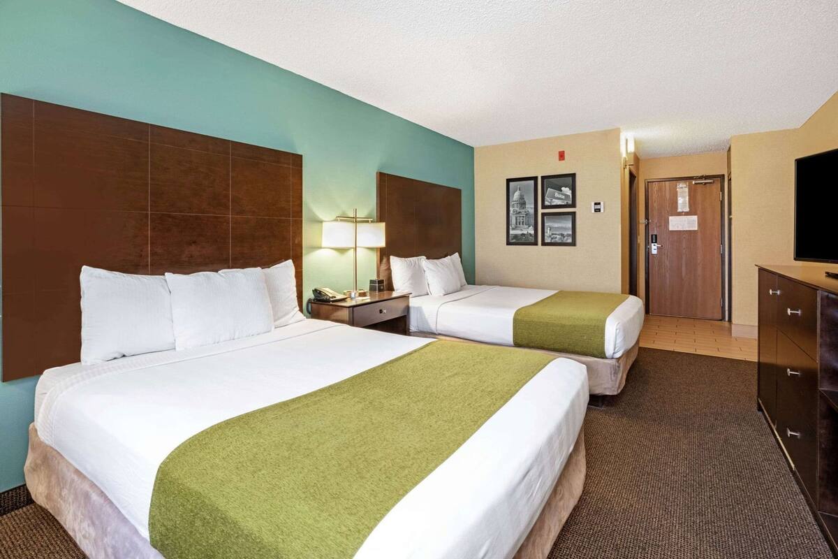 A hotel room with two beds, a desk, a lamp, a television, and a door.