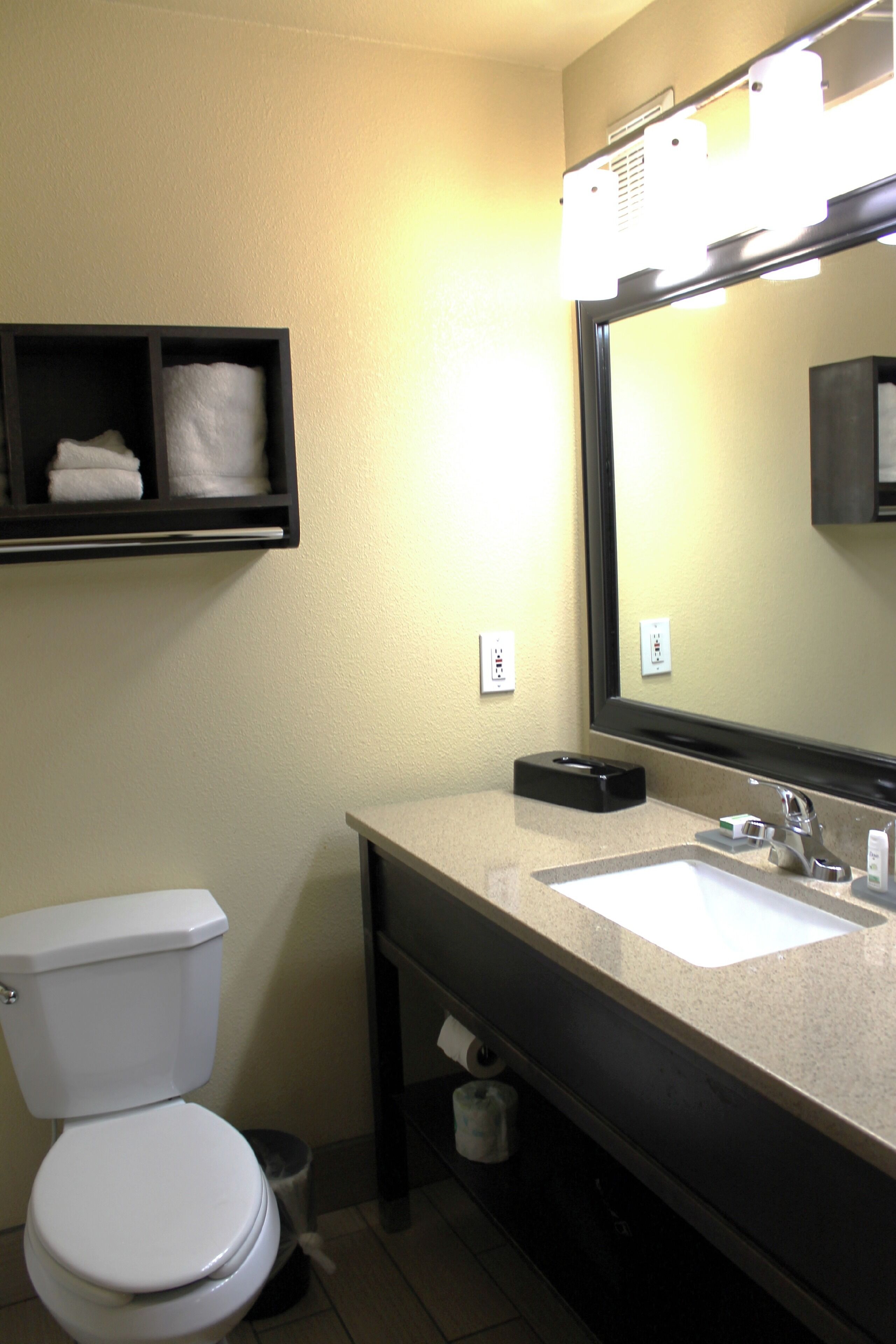 A bathroom with a toilet, sink, and mirror. There is a shelf above the toilet with folded towels.
