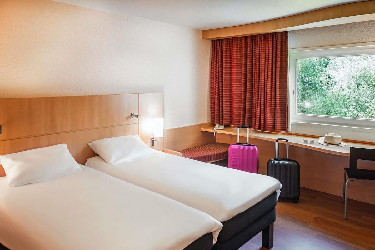 Ein Hotelzimmer mit zwei Betten, einem Schreibtisch, einem Stuhl, einem Koffer und einem Fenster mit Vorhängen.