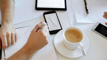 Eine Person arbeitet an einem Schreibtisch mit Unterlagen, einem Smartphone, einer Kaffeetasse und einem Tablet.